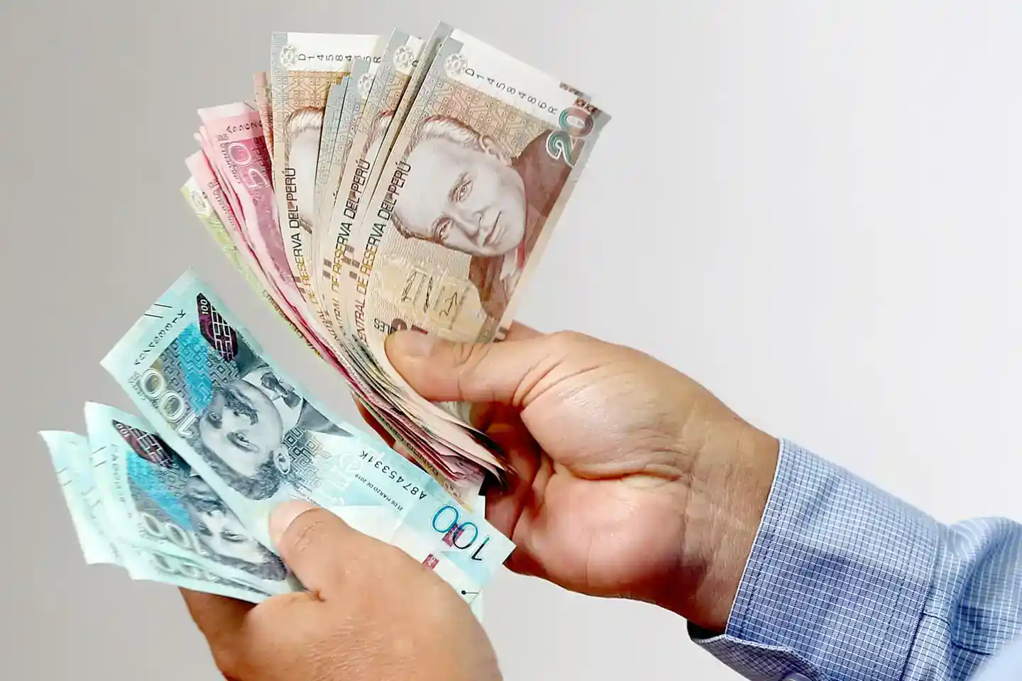 Dinero en billetes de Perú, monedas de 100 y 200 soles en manos de persona con camisa de cuadros, en fondo blanco.