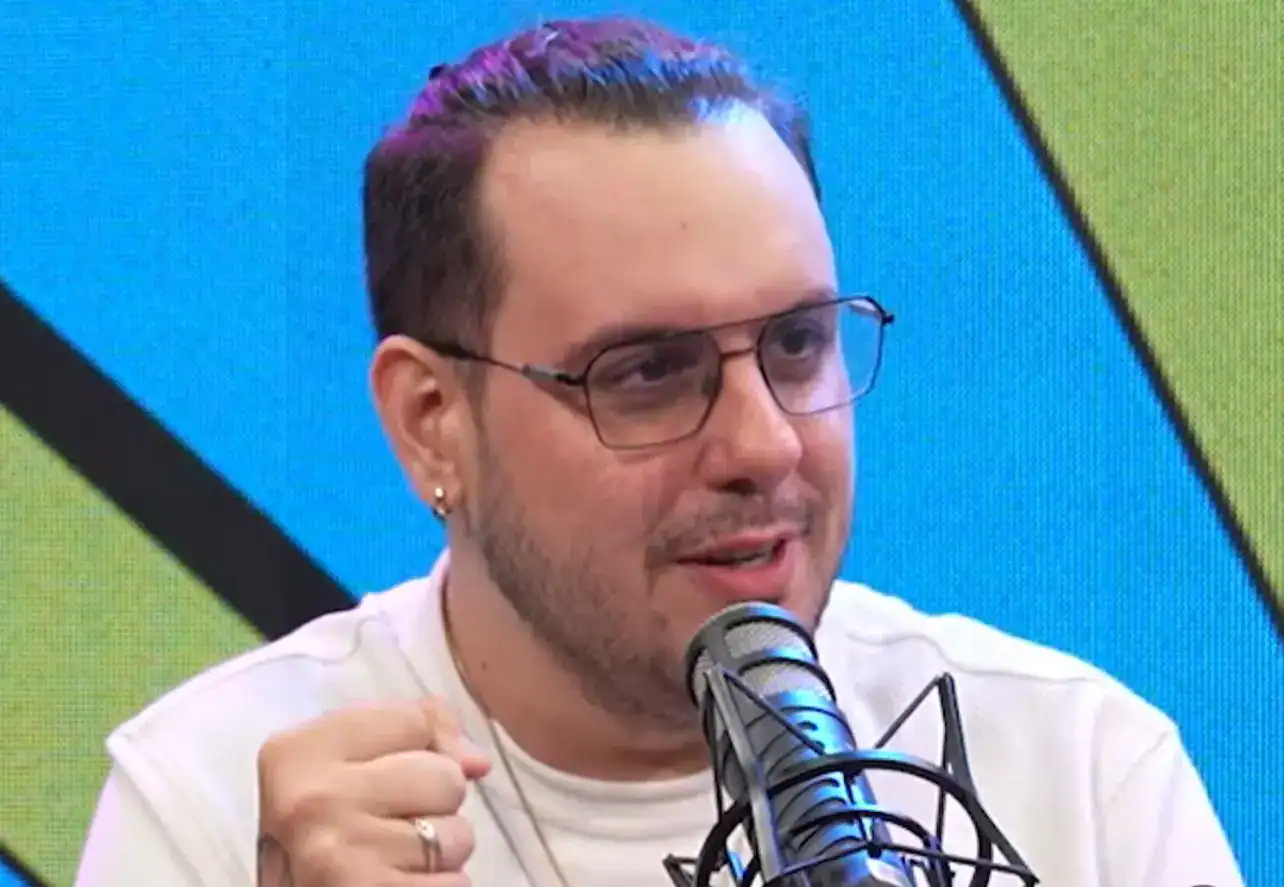 Hombre con gafas hablando en un micrófono en estudio de radio o televisión, con fondo colorido en tonos azul y verde.