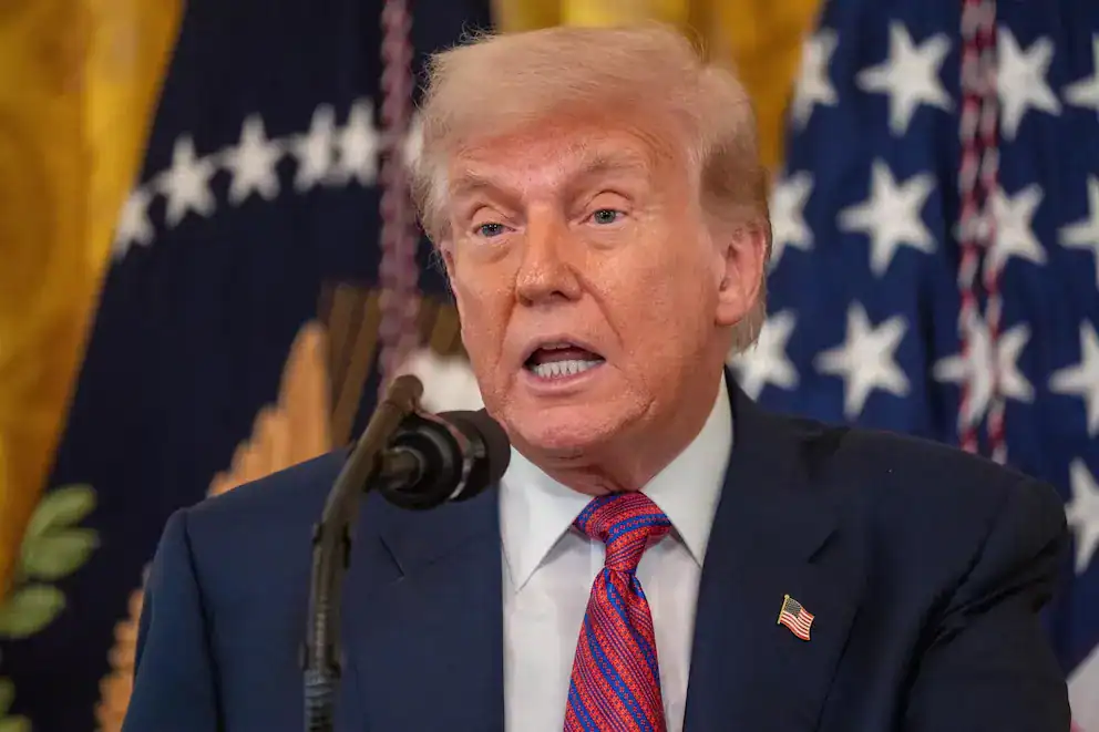 Donald Trump hablando en una conferencia, vestido con traje formal y corbata, en un ambiente oficial con banderas de EE. UU. de fondo.