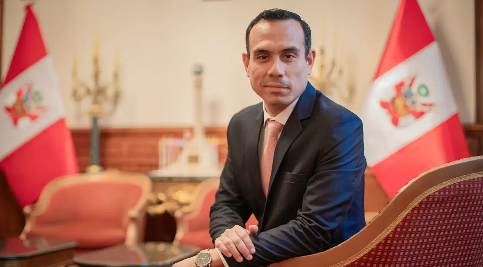 Hombre en traje formal con banderas de Perú en fondo, en una oficina gubernamental.